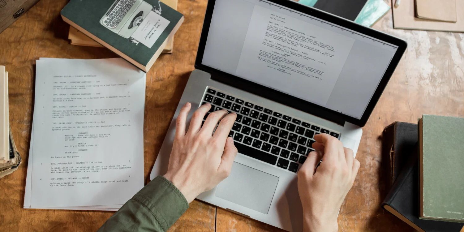 A person typing on his laptop with a printed document beside it.